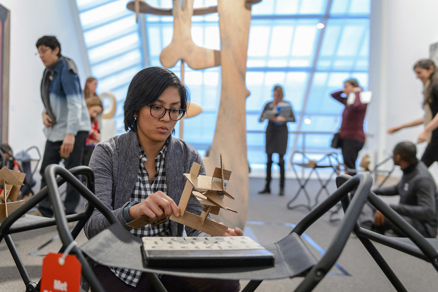 An adult educator kneels in front of a folding stool and plays with a DIY sculpture made from planes of interlocking cardboard within a skylight-filled gallery, in front of a sculpture of interlocking marble forms by Isamu Noguchi